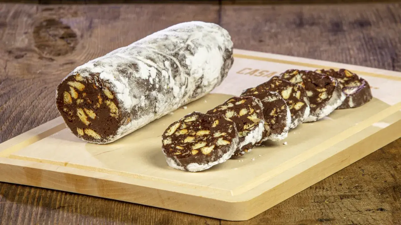 Un rotolo di salame al cioccolato, cosparso di zucchero a velo, è affettato su un tagliere di legno. Pezzi croccanti di biscotto si mescolano al cioccolato fondente.