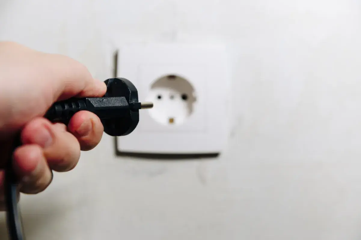 Dłoń trzyma wtyczkę, zbliżając ją do gniazdka. Uważaj, by nie doświadczyć porażenie prądem z gniazdka!