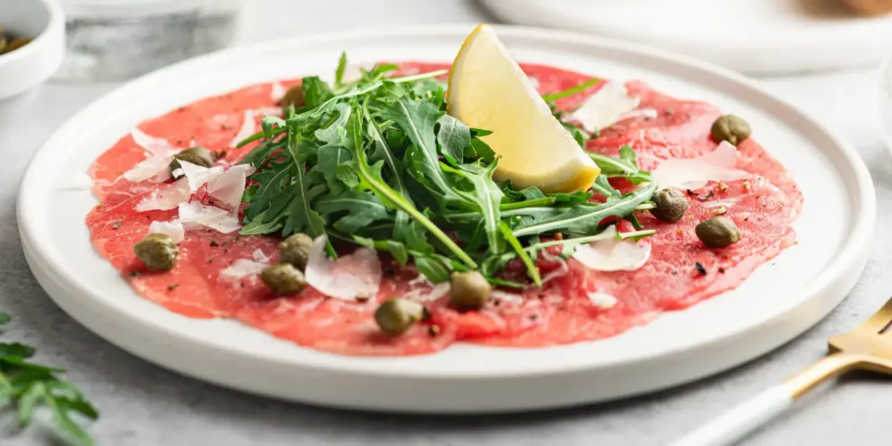 En tallrik med tunnskivad carpaccio di manzo, toppad med rucola, kapris, parmesan och en citronklyfta.