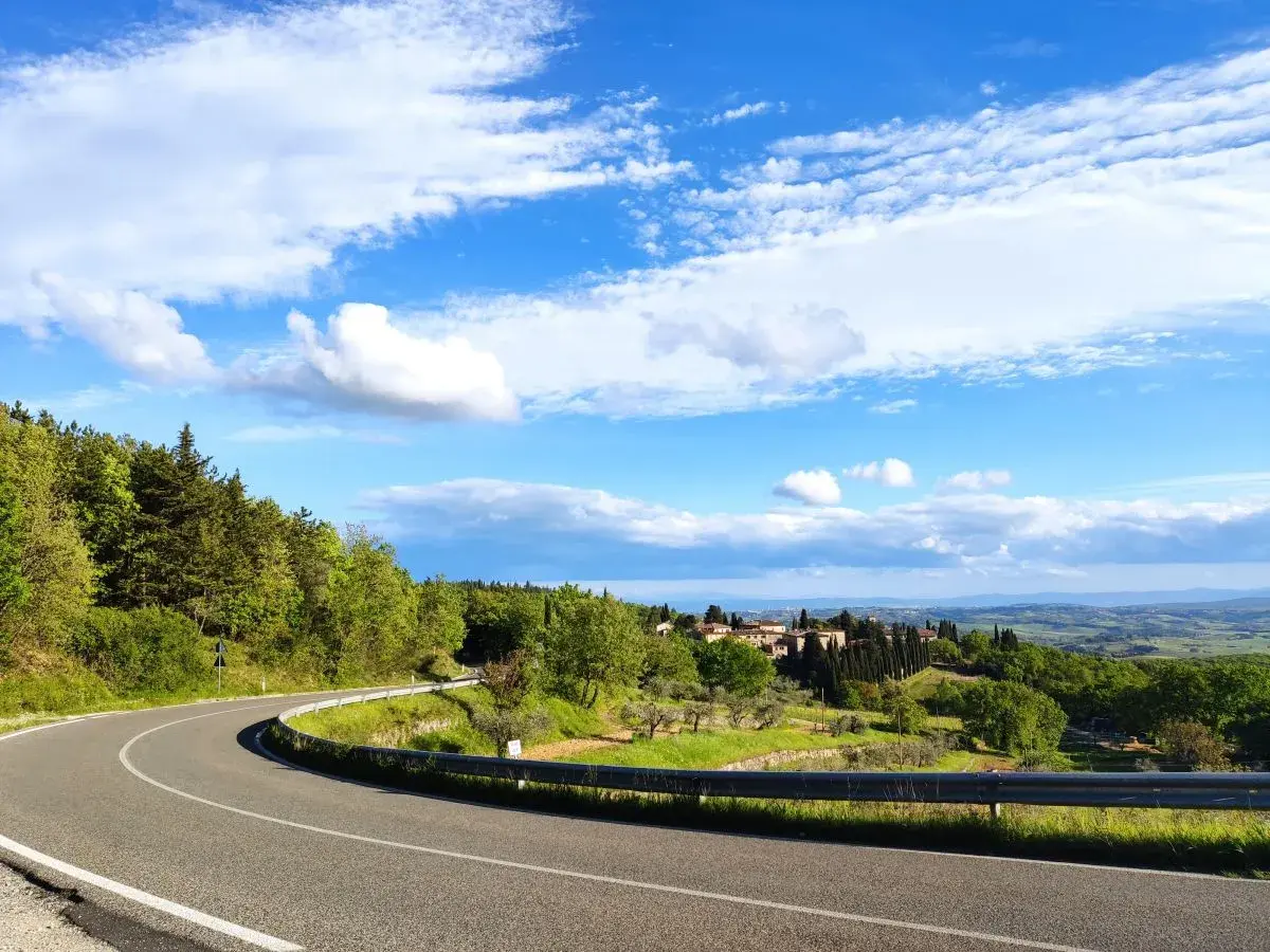 Strada SR 222 Chiantigiana serpeggia tra colline verdi e boschi, con un borgo toscano in lontananza sotto un cielo azzurro e nuvoloso.