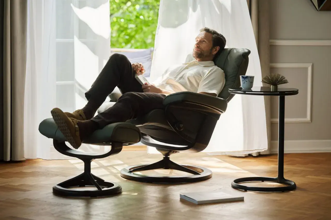 Man vilar i en bekväm Ekornes fåtölj med fotpall, njuter av ljuset från fönstret.