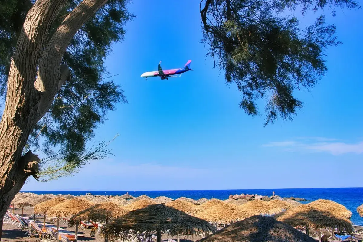 Samolot przelatuje nad plażą z parasolami, tuż przed lądowaniem na **Santorini lotnisko**.