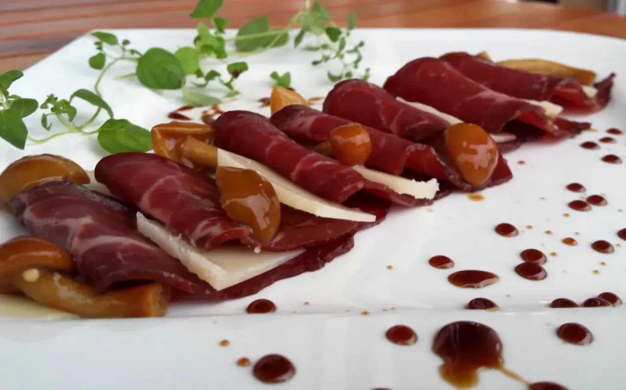 Cienkie plastry bresaoli, ser, marynowane grzyby i sos balsamiczny na białym talerzu. Tak wygląda bresaola, co to za przysmak!
