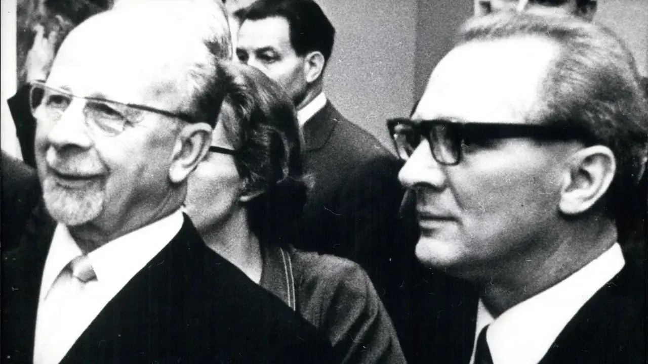 Ein Mann mit Brille und Bart, ein DDR-Chef, lächelt. Im Hintergrund weitere Personen.