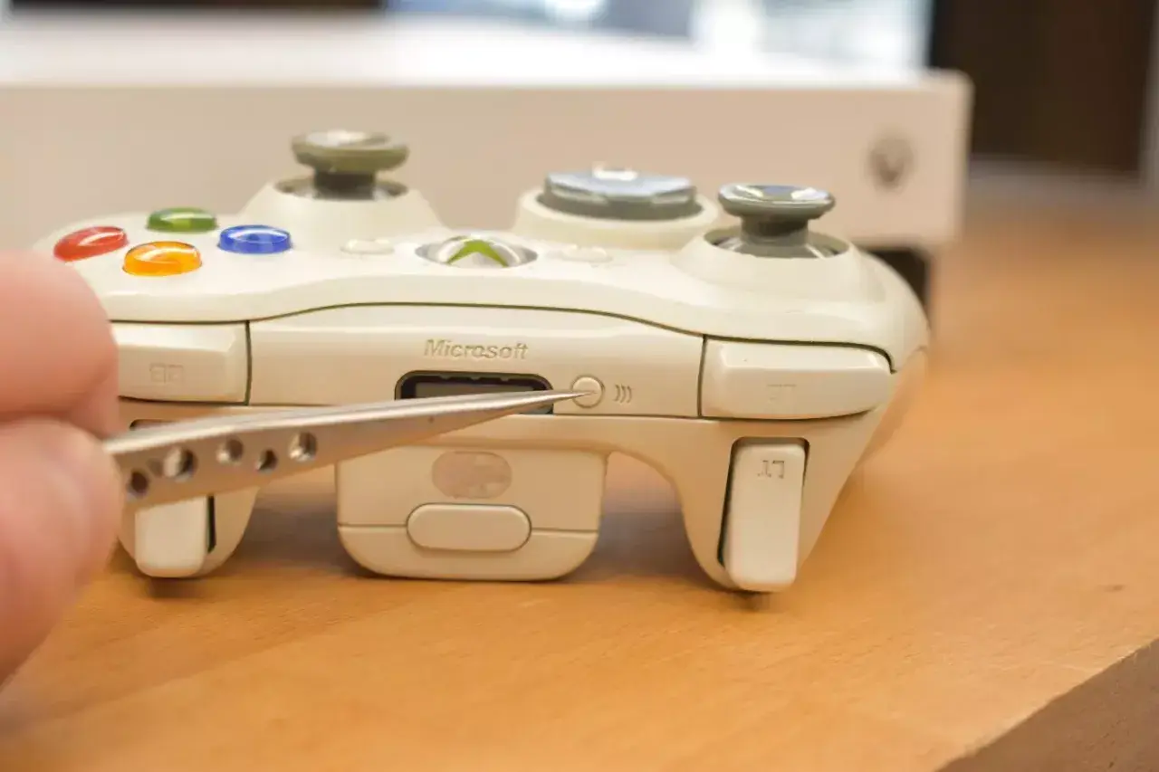 Pęseta zbliża się do przycisku synchronizacji na białym padzie Xbox. Dowiedz się, jak podłączyć drugiego pada Xbox.