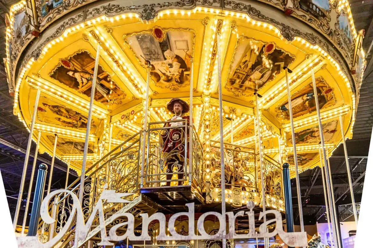 Złota karuzela w parku rozrywki Mandoria, ozdobiona malowidłami i światłami, z postacią w stroju z epoki.