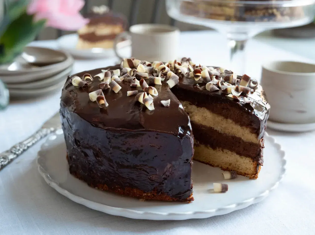 En läcker chokladtårta med blank chokladglaze till tårta, dekorerad med chokladlockar. En bit är uppskuren och visar lager av ljus och mörk fyllning.