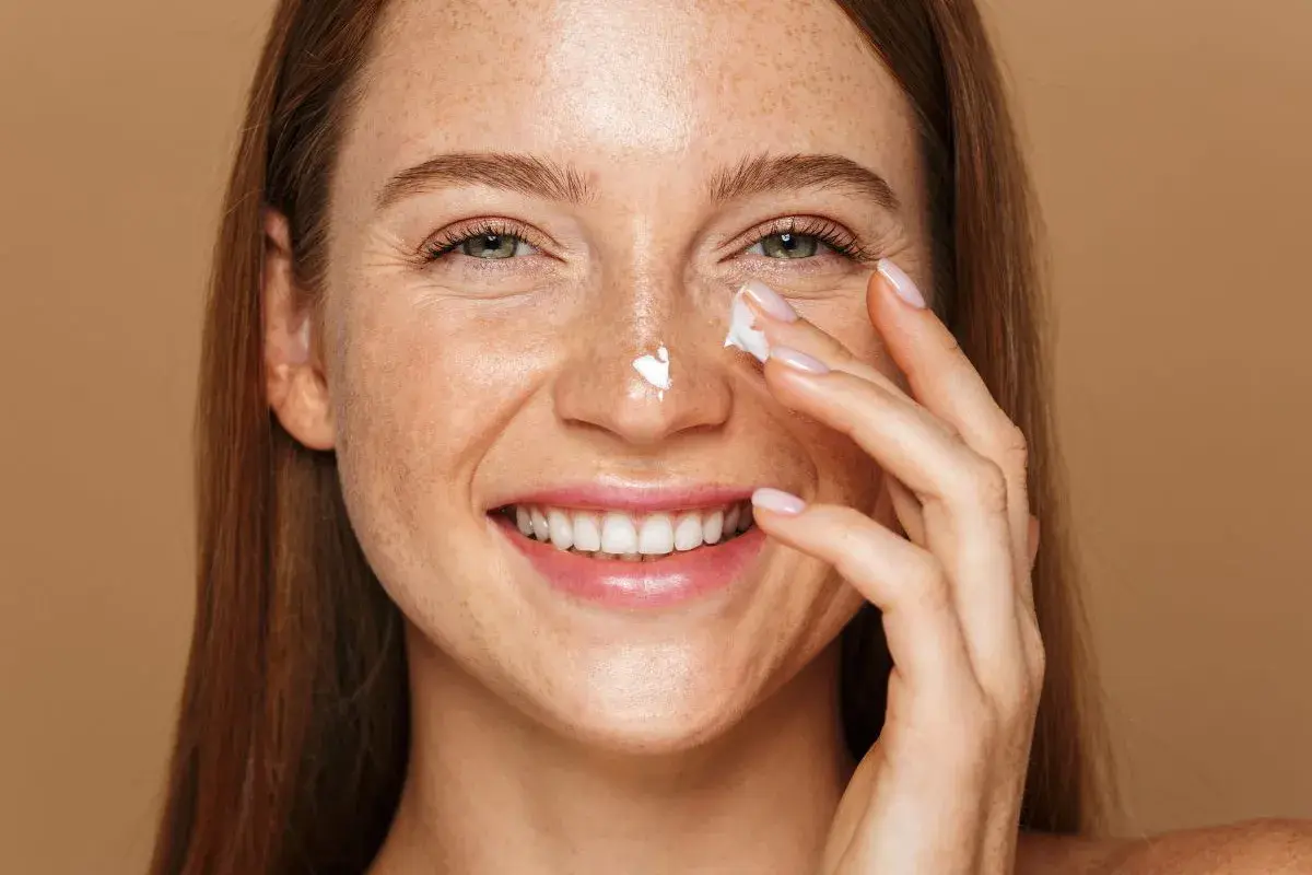Uśmiechnięta kobieta z piegami aplikuje krem na nos. Pamiętaj o ochronie przed słońcem, zwłaszcza stosując retinol.