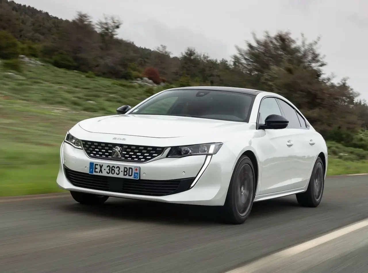 Biały Peugeot 508 jedzie po krętej drodze wśród zielonych wzgórz.