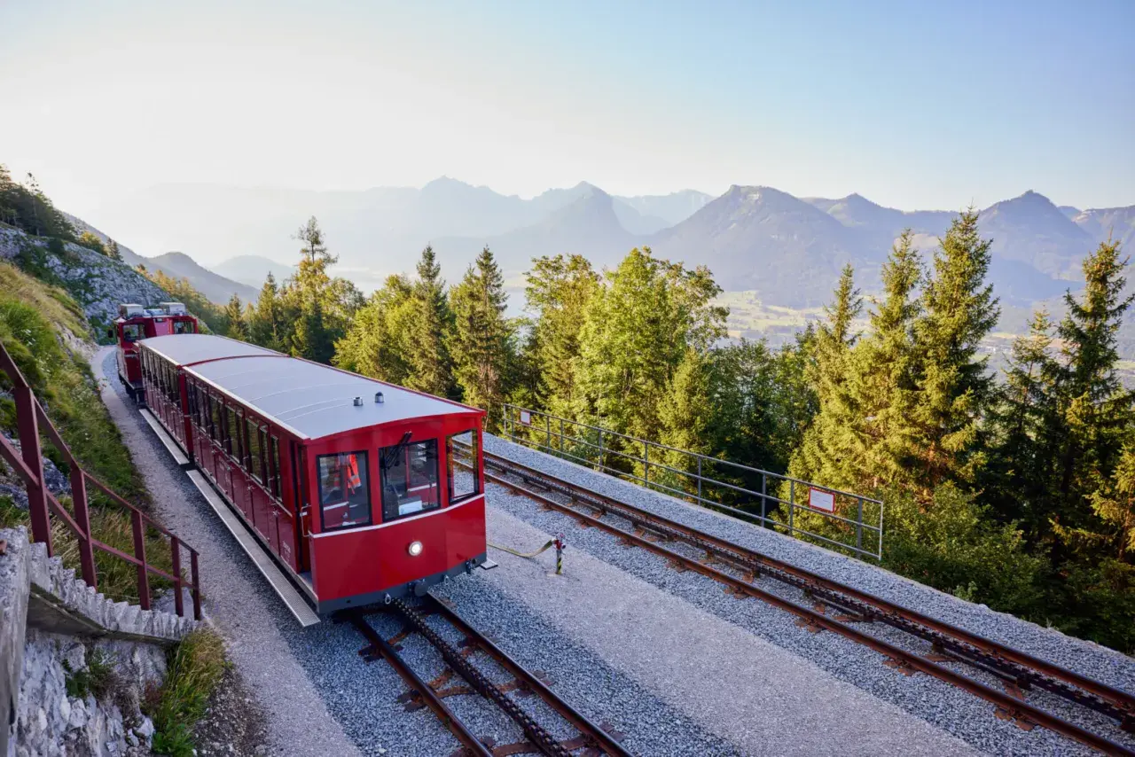Czerwony wagon kolejki zębatej jedzie po torach w górę zbocza. W tle góry i lasy.