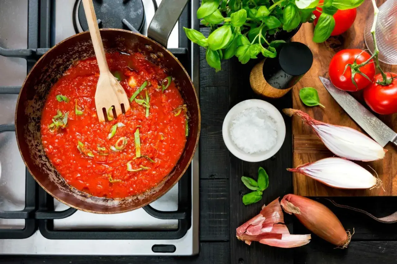 En kastrull med bubblande tomatsås och en träsked. Ingredienser för ett tomatsås recept: färska tomater, schalottenlök, basilika och salt.