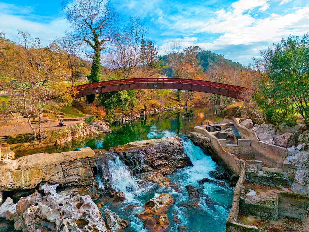Cascada y puente de madera en la vía verde Puente Viesgo, rodeados de árboles otoñales y un cielo azul vibrante.