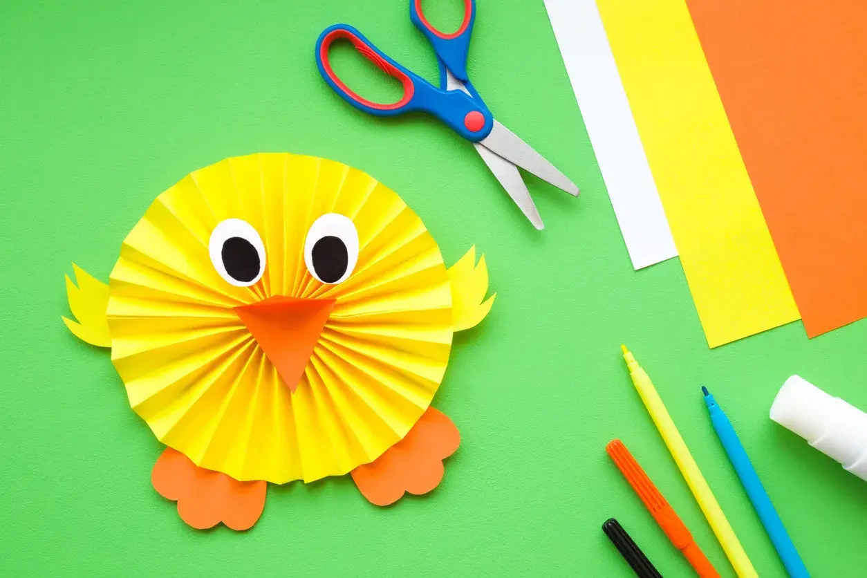 Un pollito de papel, ideal para manualidades fáciles para niños de 3 a 6 años. Tijeras, rotuladores y cartulinas de colores completan la escena.