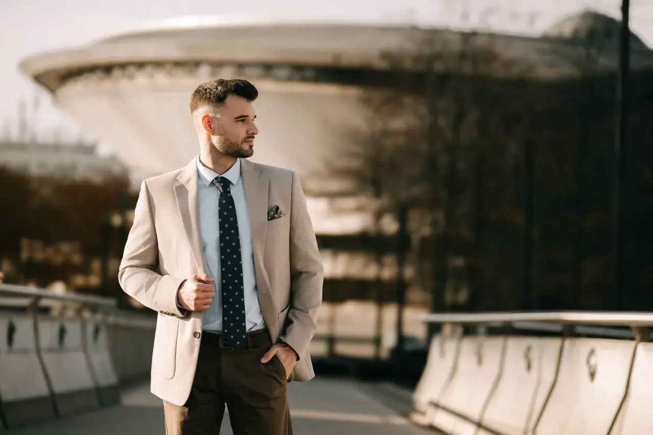 Elegancki mężczyzna w beżowej marynarce, na który guzik zapina się marynarkę, stoi na tle nowoczesnej architektury.