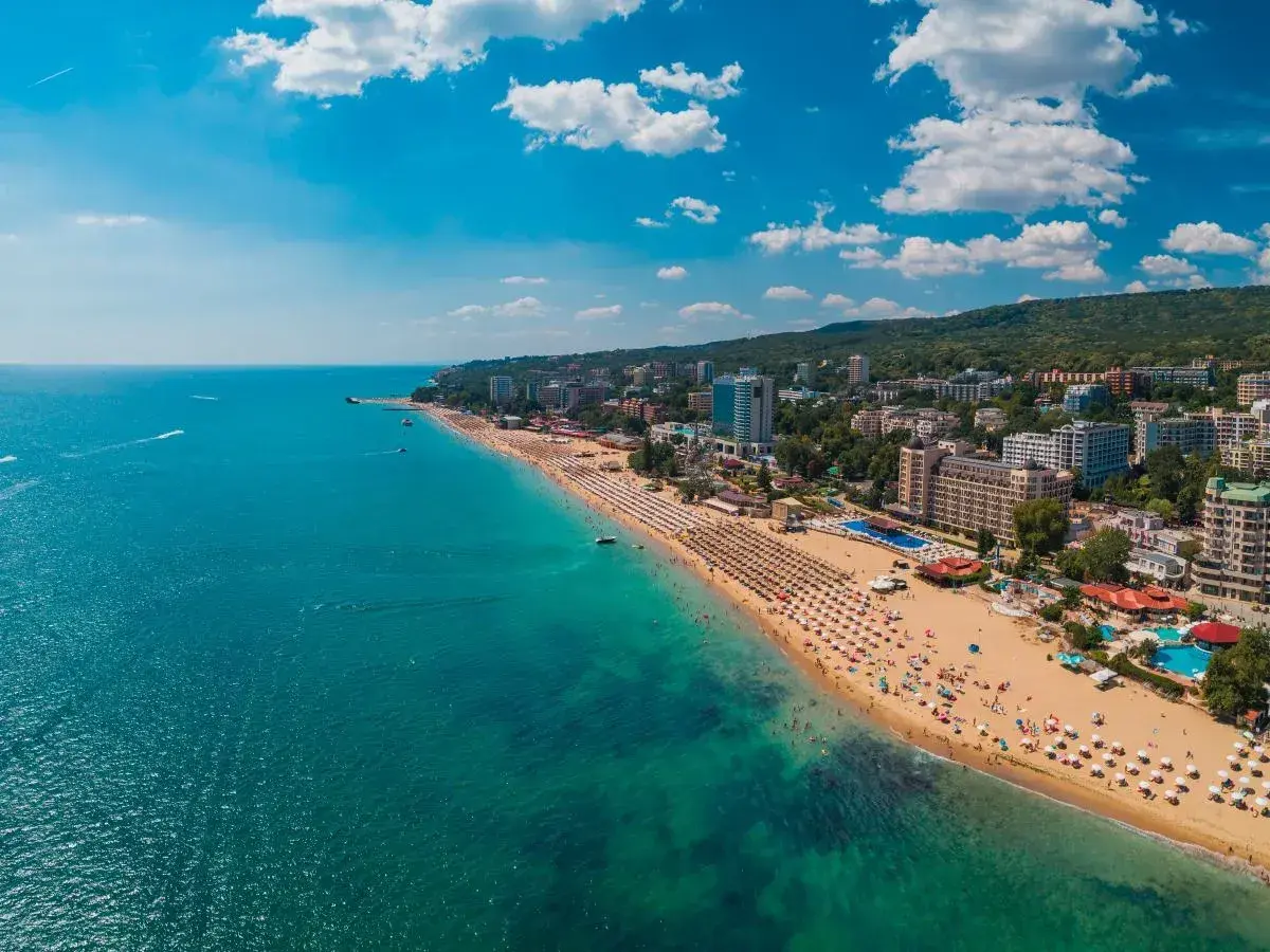 Słoneczna plaża z mnóstwem leżaków i parasoli, tętniące życiem kurorty nad morzem, a w oddali zielone wzgórza. Idealne miejsce na wakacje.