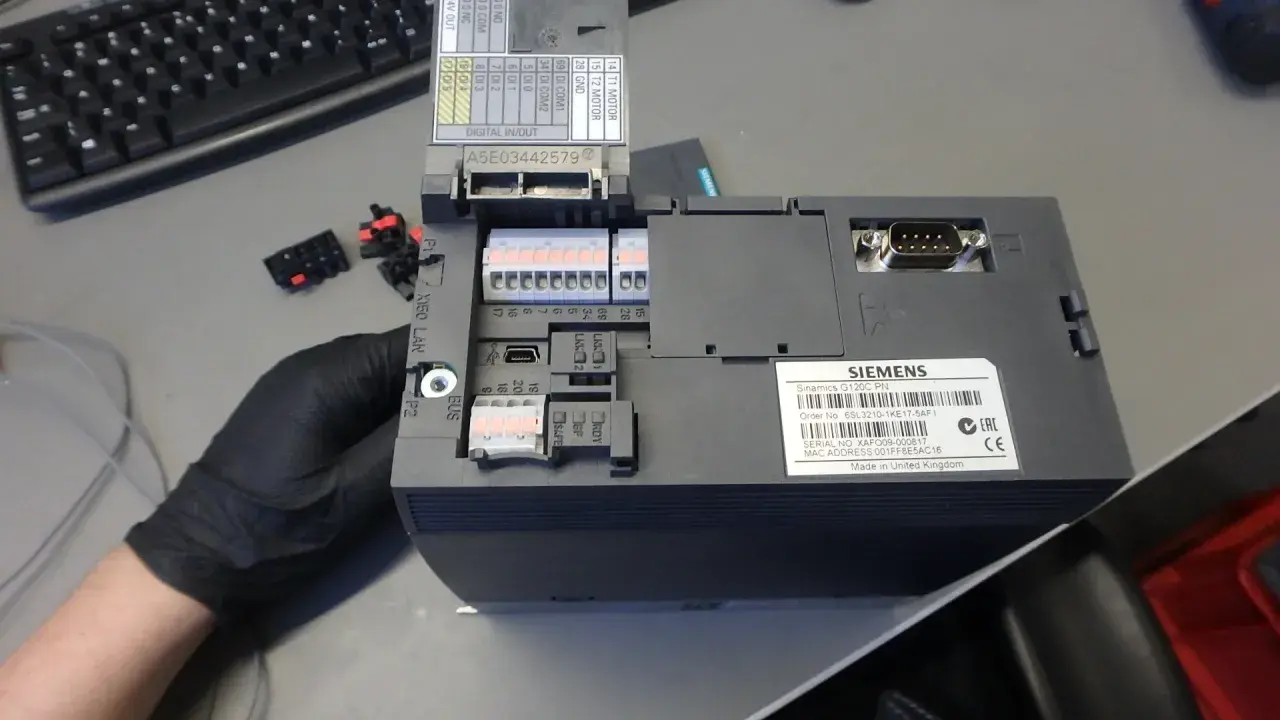 Ręka w czarnej rękawiczce trzyma sterowniki Siemens Sinamics G120C PN. Widać złącza, porty USB i etykietę z numerem seryjnym.