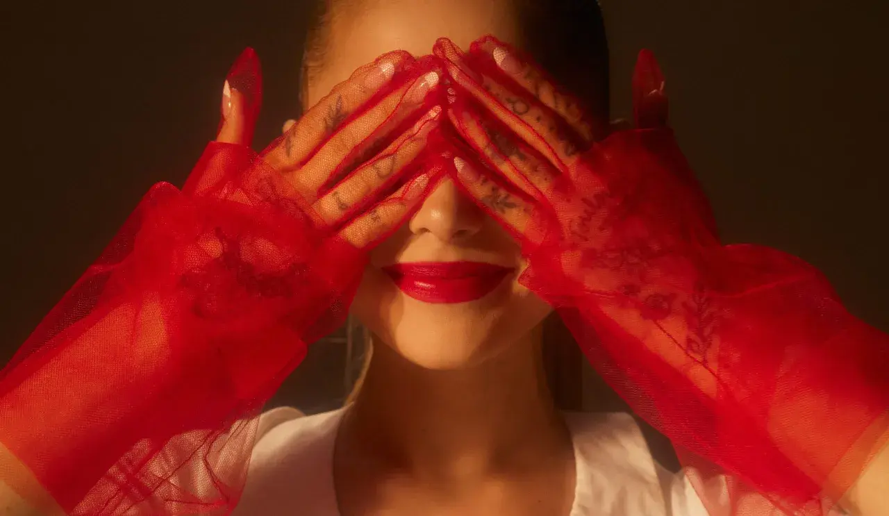 Ariana Grande 2024 w czerwonych rękawiczkach zakrywa oczy, odsłaniając usta i tatuaże na dłoniach.