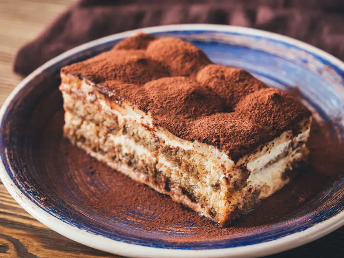Une part de tiramisu, saupoudrée de cacao, sur une assiette bleue. Un délice qui évoque tous les ingrédients du tiramisu.