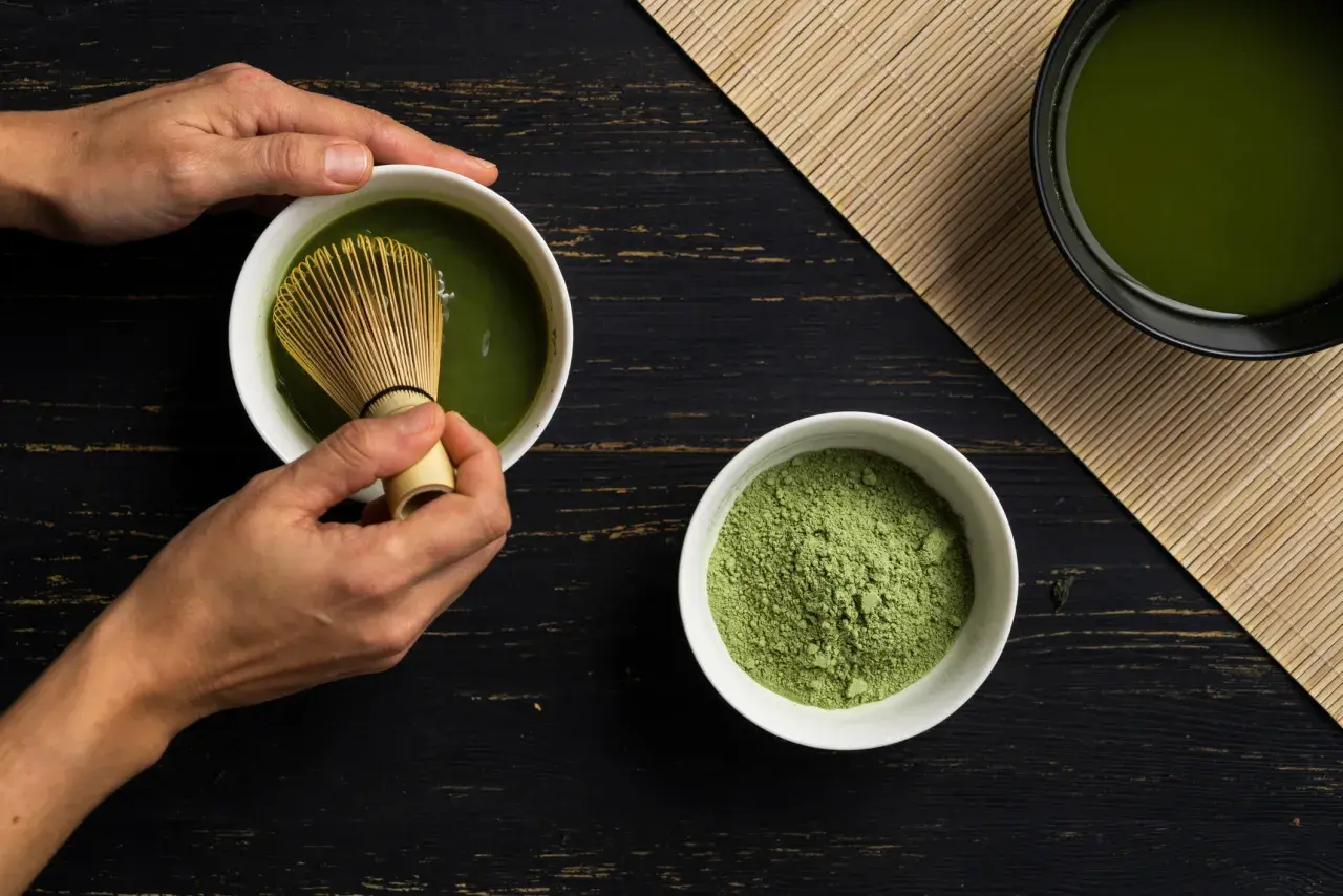 Hände bereiten Matcha zu. Pulver und fertiger Tee auf dunklem Holz. Eine positive matcha tee abnehmen erfahrung beginnt hier.