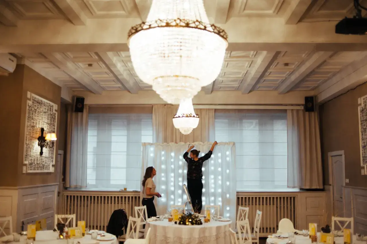 Dwie kobiety przygotowują salę weselną, jedna stoi na drabinie, dekorując tło światełkami. To początek drogi, jak zostać wedding plannerką.