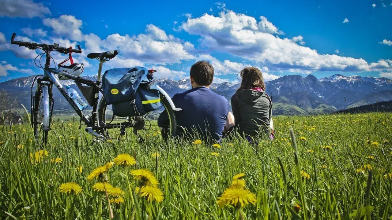 Para na rowerach odpoczywa na łące pełnej mleczy, podziwiając Tatry. To idealne miejsce na szlak papieski Gubałówka.