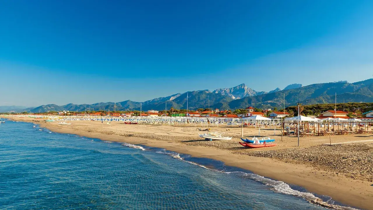 Słoneczna plaża w Toskanii, z rzędami białych parasoli i leżaków, w tle góry i błękitne morze.