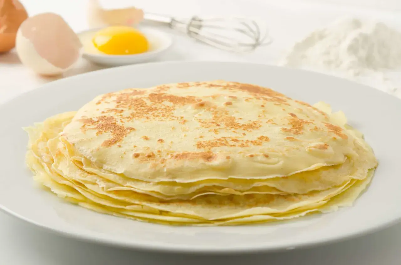 Ein Stapel goldbrauner Palatschinken auf einem Teller, daneben Eier, ein Schneebesen und Mehl. Perfekt für ein Palatschinken Rezept!