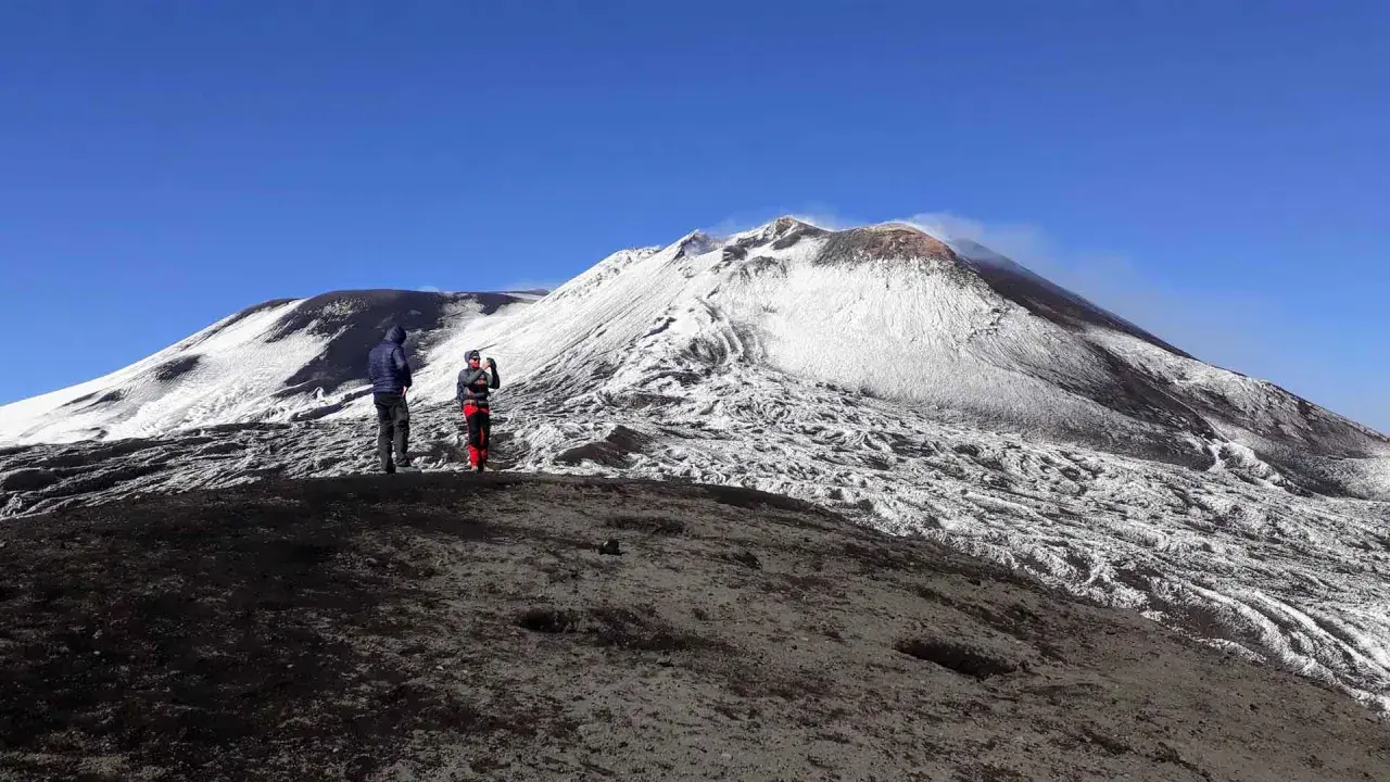 Dwie osoby stoją na zboczu wulkanu Etna pokrytego śniegiem. W tle widać dymiący krater.