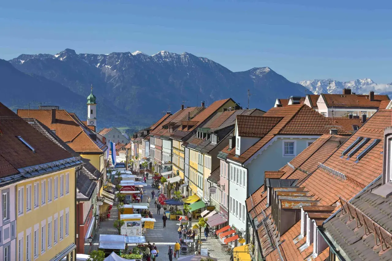 Bunte Häuserfronten säumen eine belebte Straße mit Marktständen, im Hintergrund majestätische Berge. Entdecke Murnau Geheimtipps!