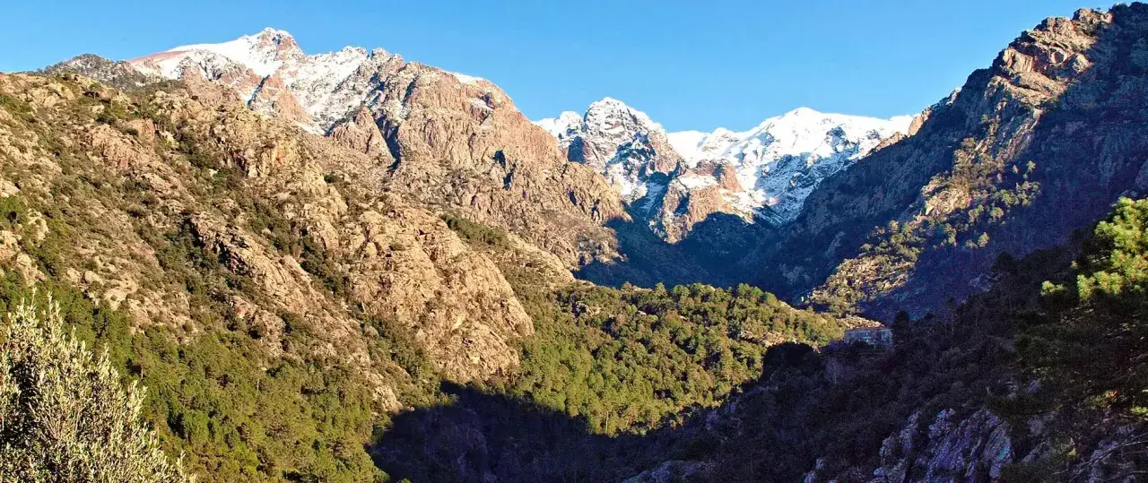 Vue spectaculaire de **mare e monti** : des montagnes enneigées surplombent une vallée verdoyante, avec un soleil éclatant illuminant le paysage.