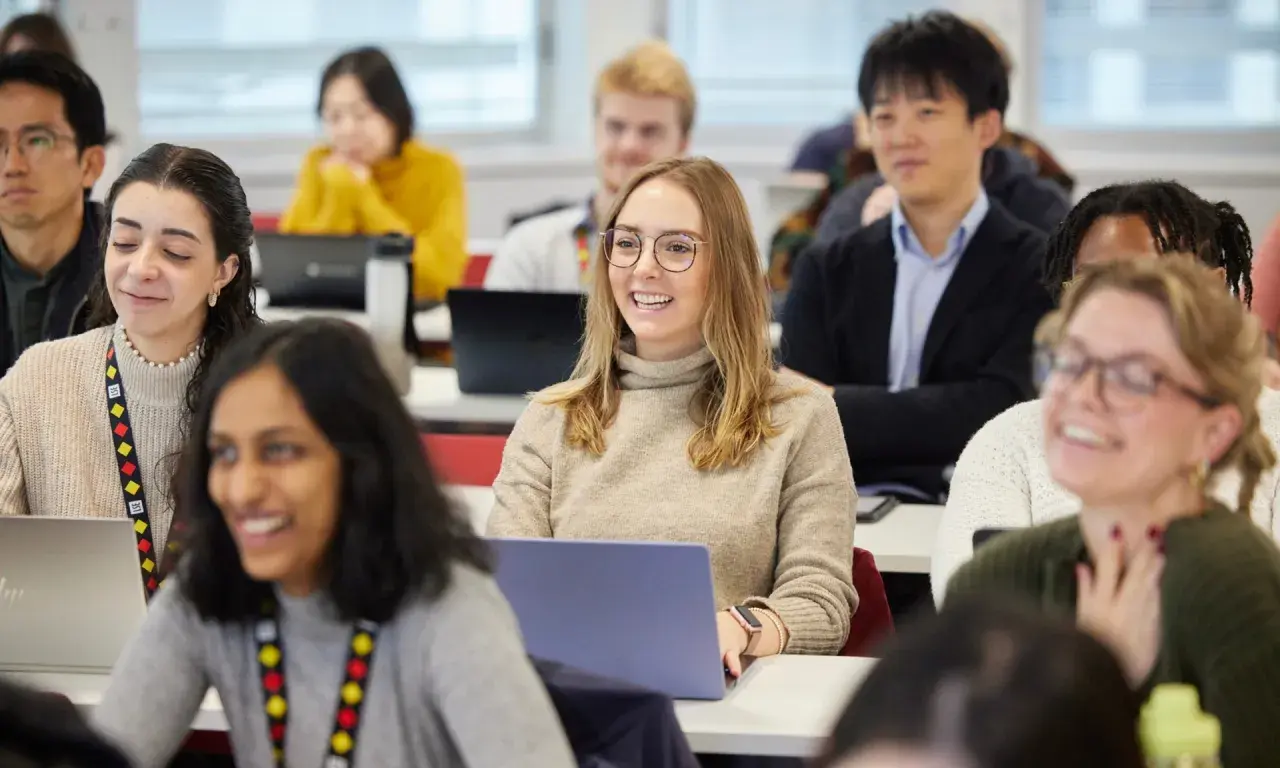 Students in a classroom, some with laptops, are engaged in a lesson. One student smiles, perhaps anticipating her MPA credential.