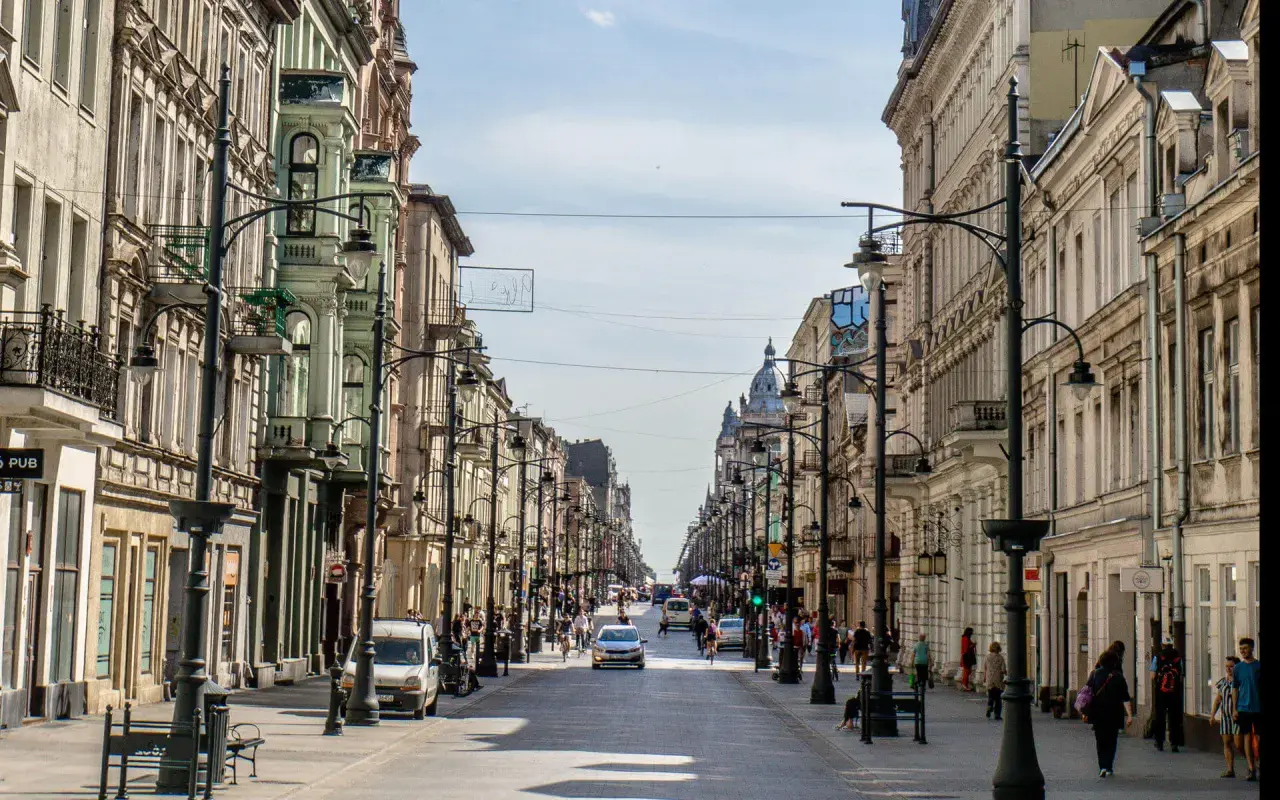 Belebte Straße in **Łódź, Polen**, gesäumt von historischen Gebäuden und Straßenlaternen. Autos und Fußgänger beleben die Szene.
