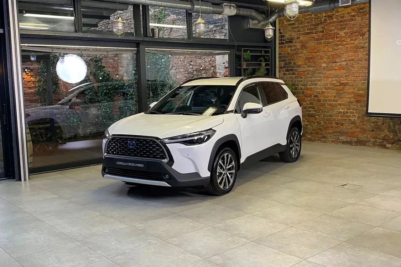 Biała Toyota Corolla Cross Hybrid stoi w nowoczesnym wnętrzu z ceglaną ścianą i dużymi oknami.