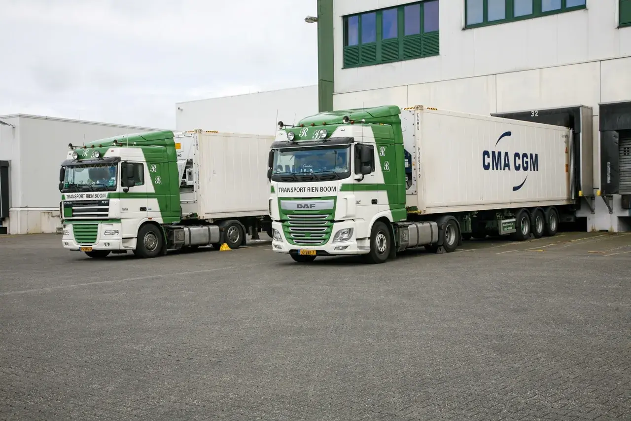 Zwei weiße und grüne LKWs stehen bereit, um entladen zu werden. Einer trägt die Aufschrift "Transport Rien Boom", der andere "CMA CGM".