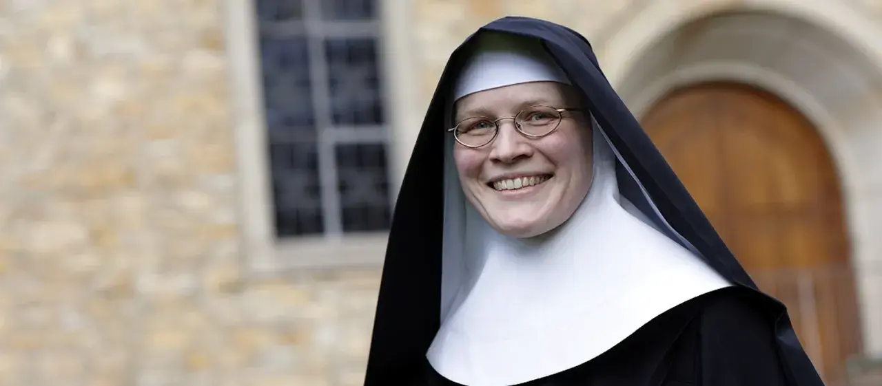 Lächelnde Nonne mit Brille vor einem Kloster. Sie hat sich entschieden, ins Kloster zu gehen und strahlt Zufriedenheit aus.