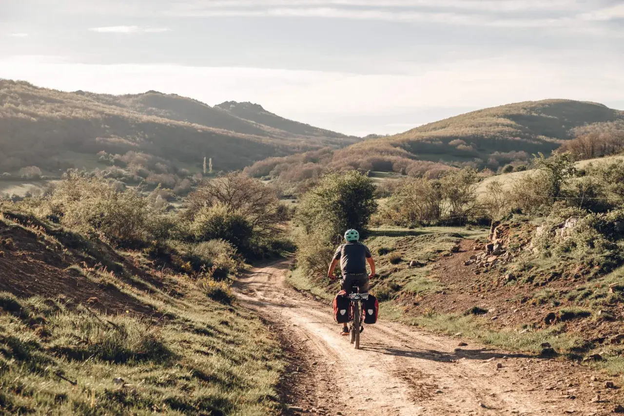 Ciclista pedaleando por un camino rural en España, disfrutando de sus viajes en bicicleta por España. Paisaje montañoso y vegetación verde.