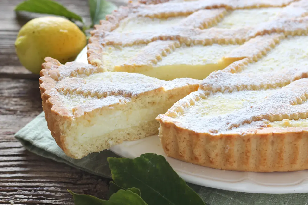 Una fetta di crostata di ricotta, cosparsa di zucchero a velo, accanto a un limone e foglie verdi su un tavolo di legno.