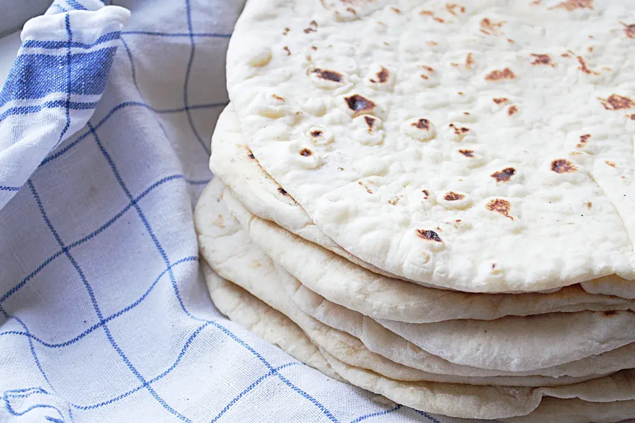 En hög med hemgjorda tortillas, perfekta för ditt nästa tortilla recept. En blårutig kökshandduk ligger bredvid.