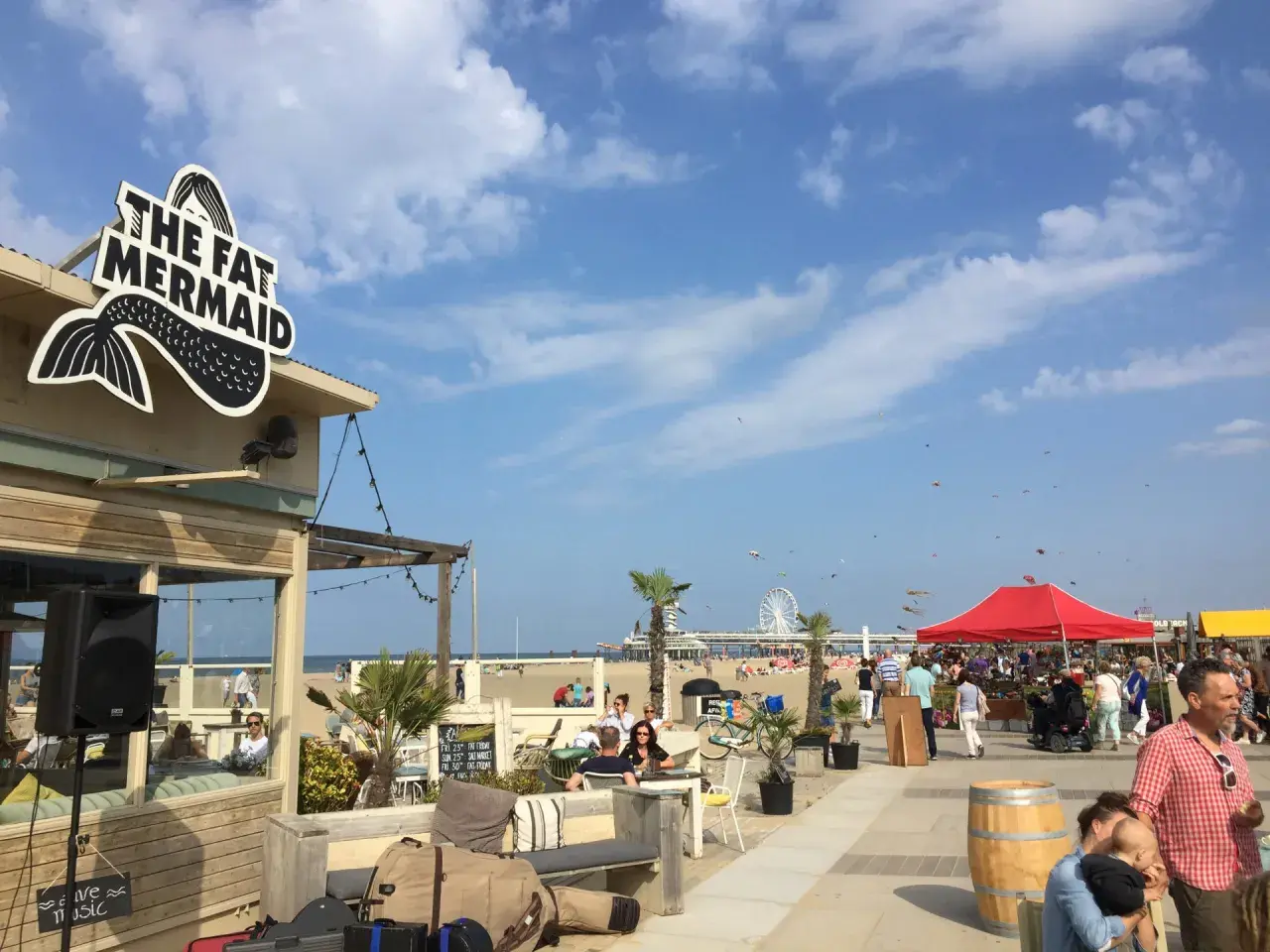 Strandpromenade in Den Haag mit dem Restaurant "The Fat Mermaid", Menschen, Sonnenschirmen und einem Riesenrad. Tolle Den Haag Unternehmungen!