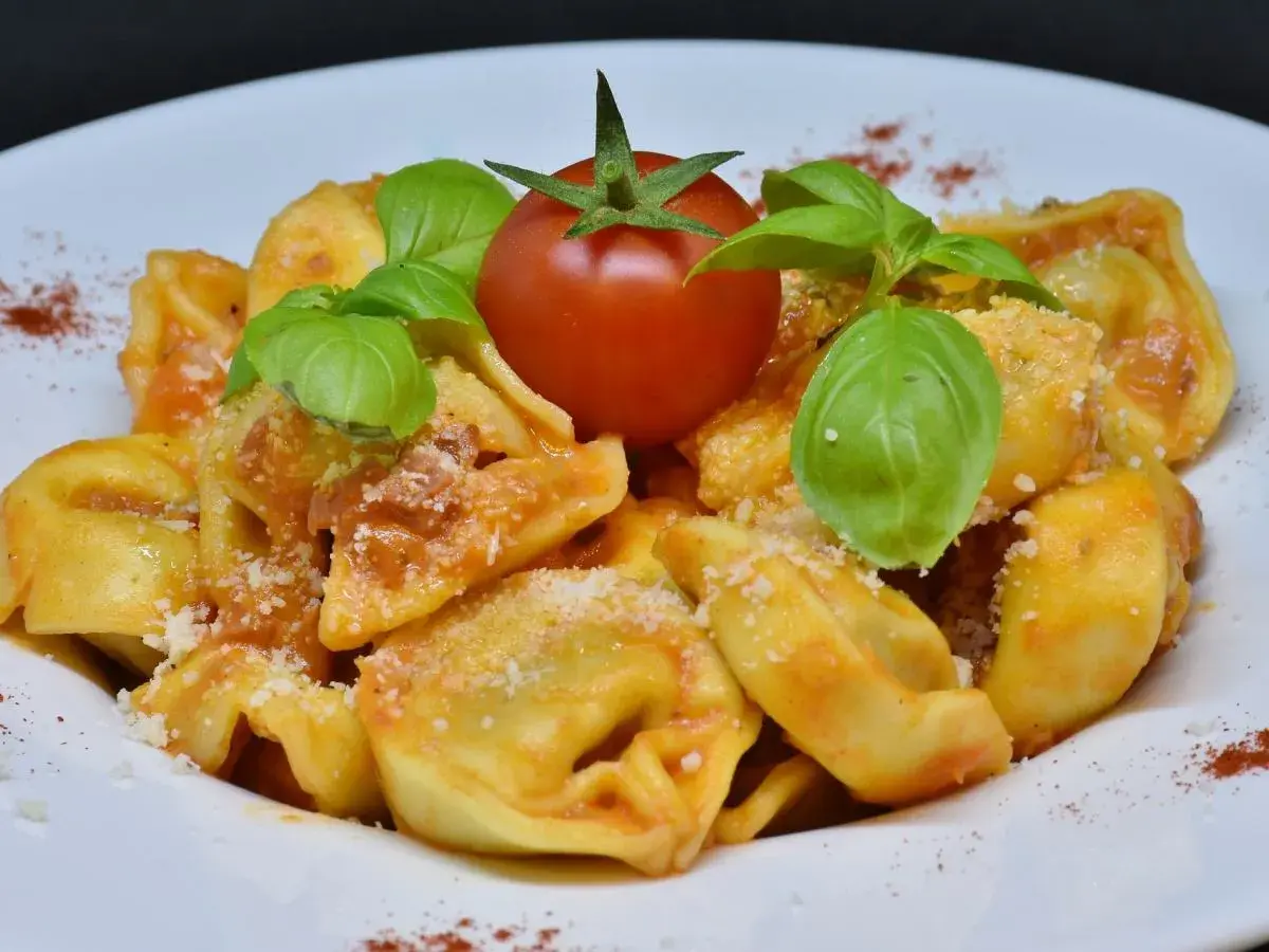 Pyszne tortellini na talerzu, polane sosem pomidorowym, ozdobione listkami bazylii i pomidorkiem.
