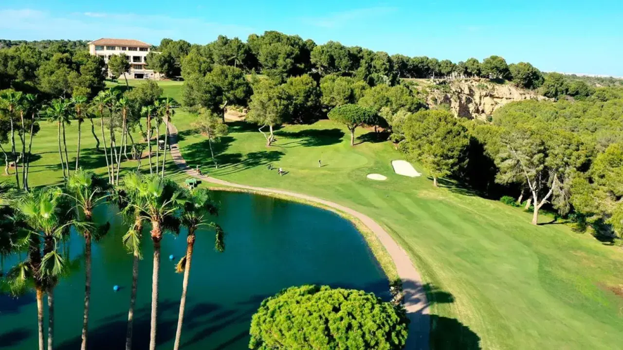 Un hermoso campo de golf con un lago azul, palmeras y un edificio al fondo. El sol ilumina el verde impecable de las ramblas golf.