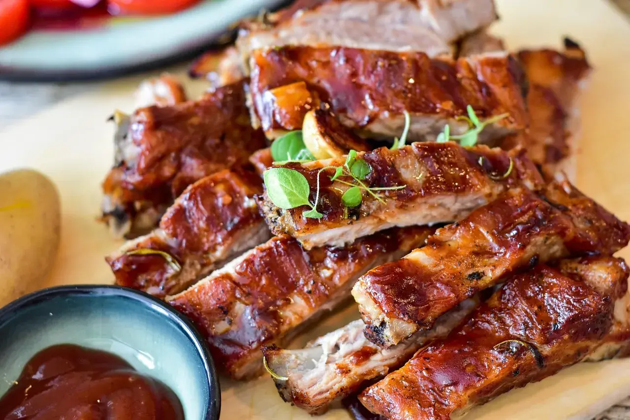 Saftige Rippchen, mariniert zum Grillen, glänzen in einer süßen BBQ-Sauce. Daneben eine Schale mit Sauce und eine Kartoffel.