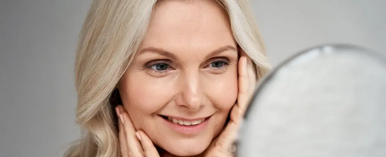 Uśmiechnięta 40-latka z blond włosami, patrząca w lustro. Delikatny makijaż na wesele podkreśla jej urodę.