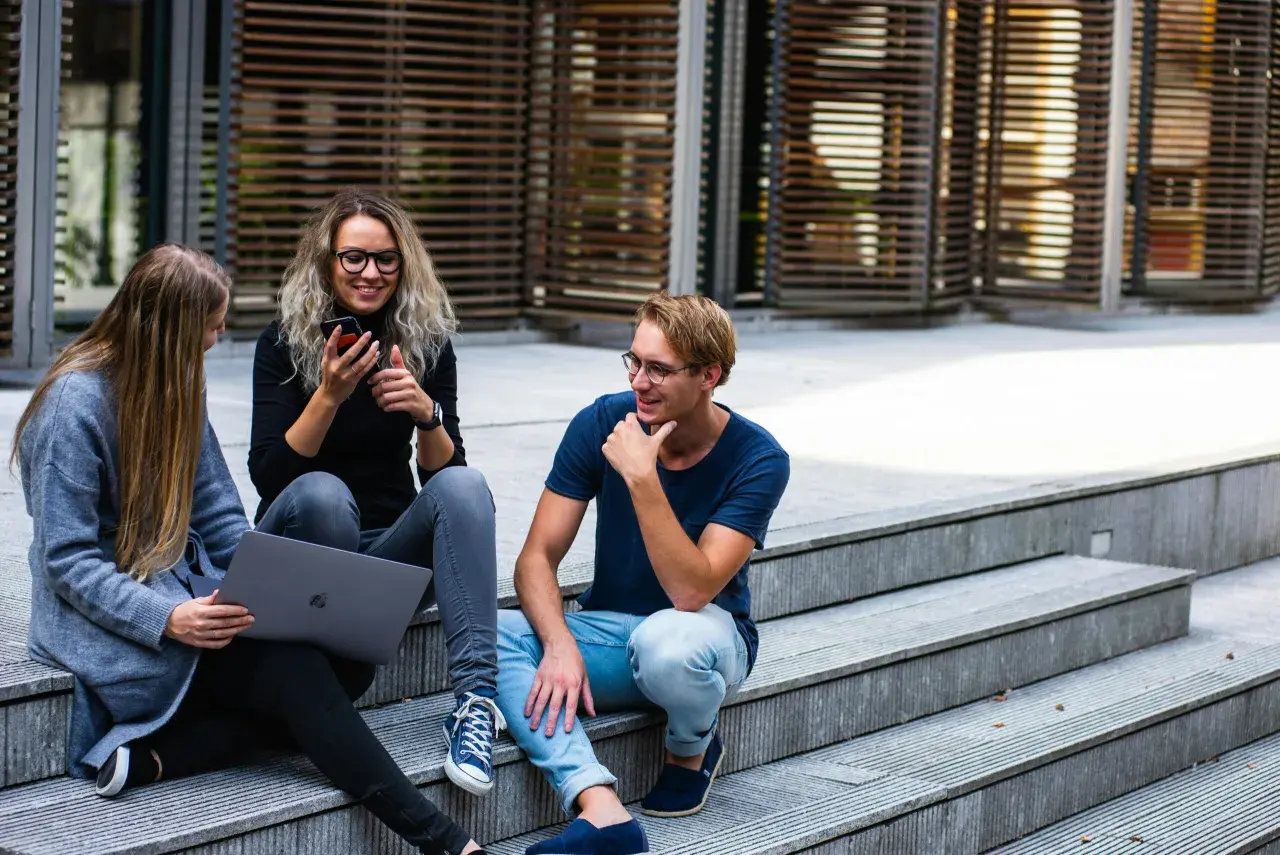 Drei junge Leute sitzen auf einer Treppe. Eine Frau mit Laptop und eine Freundin mit Handy, die sich über das Studium zuhause unterhalten.
