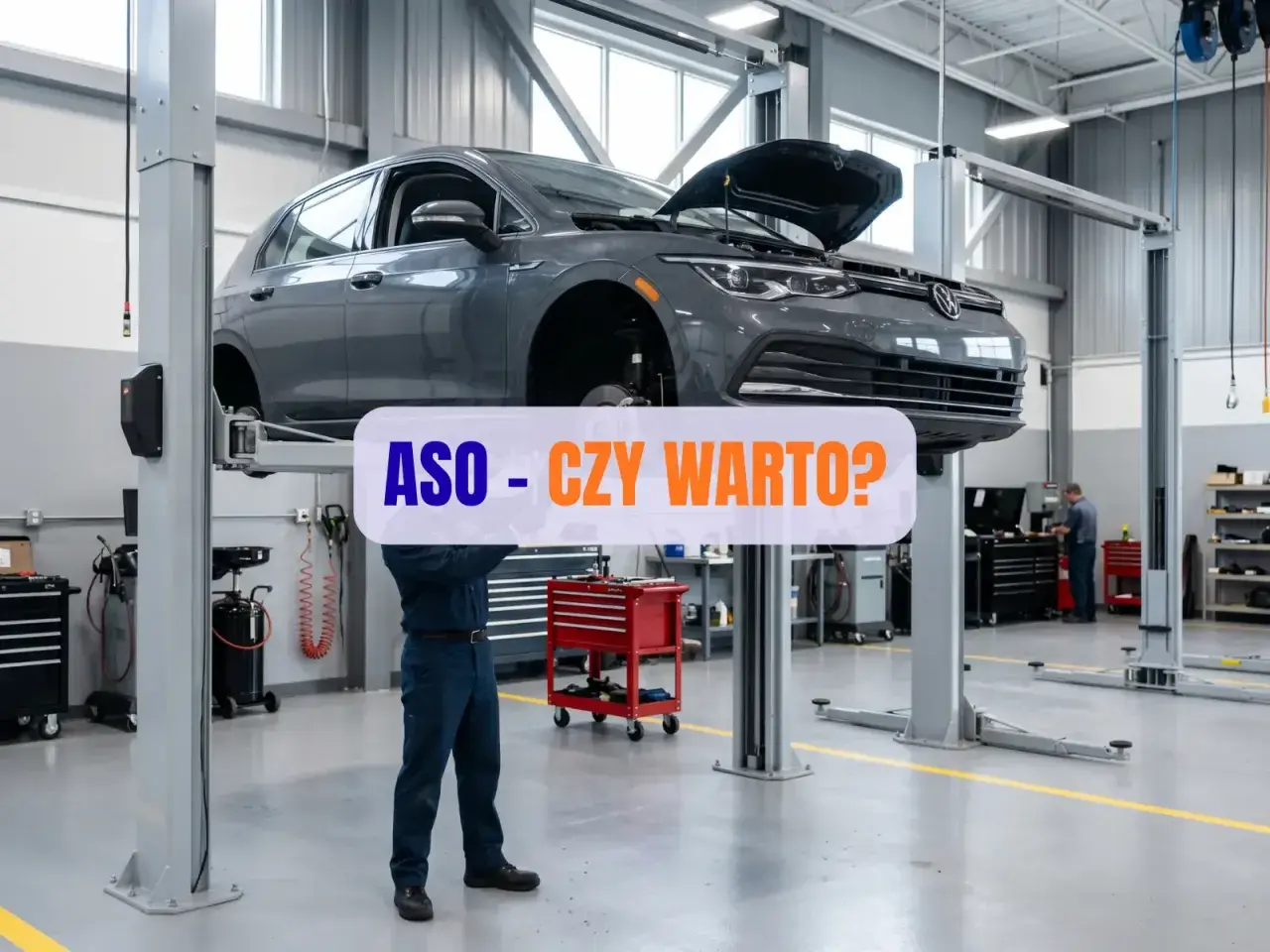 Szary samochód na podnośniku w ASO. Mechanik pracuje pod autem, a nad nim napis "ASO - CZY WARTO?".