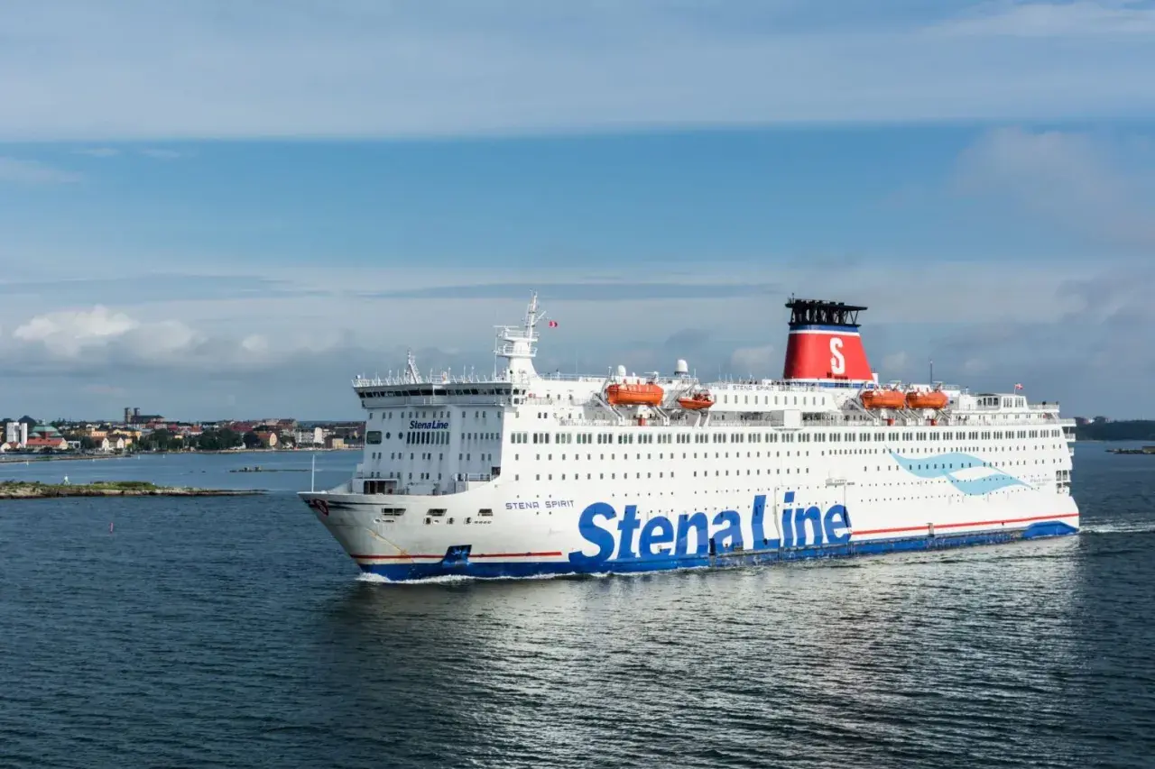 Statek Stena Line płynie po morzu, gotowy na rejs 24h. W tle widać wybrzeże z zabudowaniami.