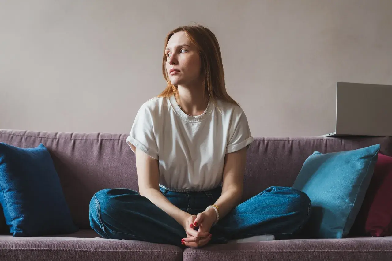 A young woman with red hair sits cross-legged on a couch, looking away thoughtfully.