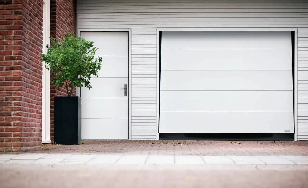 Modernt vipport garage med en vit port och en sidodörr. En grön buske i en svart kruka står bredvid.