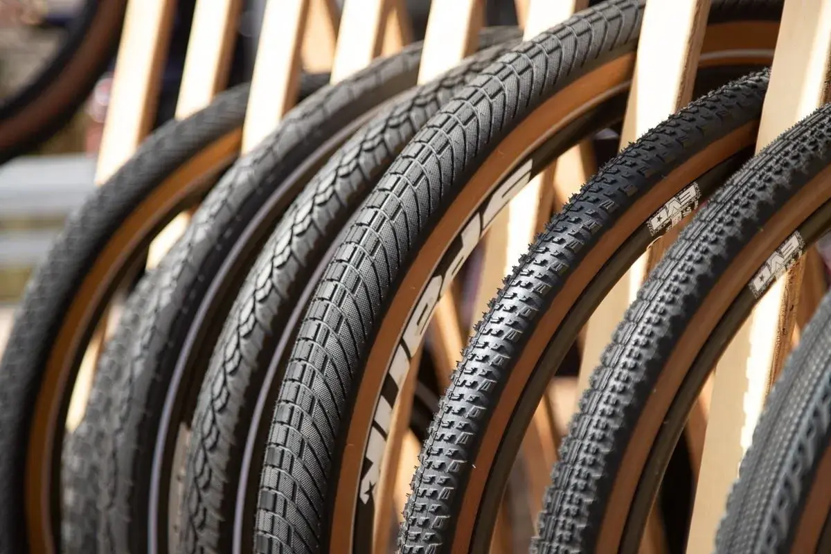 A row of diverse bicycle tires, featuring knobby mountain bike tires and smoother road bike tires, all with distinctive tan sidewalls, are displayed.