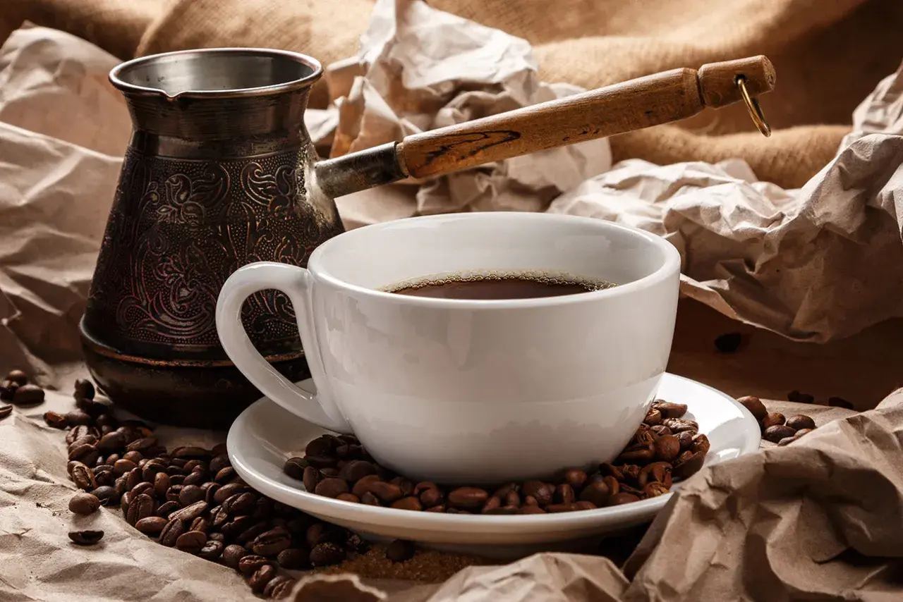 Ein traditioneller Mokkakanne und eine Tasse mit griechischem Mokka, umgeben von Kaffeebohnen und zerknülltem Papier.
